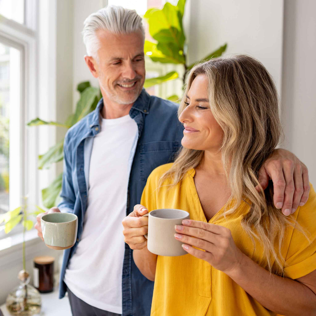Man and woman holding coffee mugs indoors with plants in the background